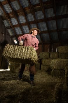 Man wearing a cowboy hat and carrying bales of hay in the barn. Vertical shot.