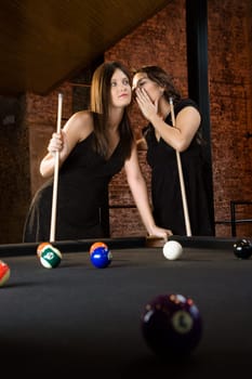 Two attractive young women at pool table as one whispers into other's ear.