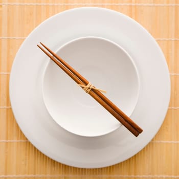 Overhead view of chopsticks lying across an empty bowl that is sitting on a plate on a bamboo mat. The dishes are white. Square shot.