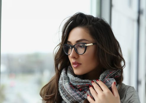 Portrait of young beautiful woman in scarf, warming hands, pensive look away, view from window. Brunette lady in glasses, mysterious look. Beauty concept