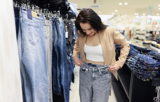 Portrait of young woman shopaholic choosing jeans in luxury store, woman try on pants on big sale in shop. Purchase, shopping, discount, addiction concept