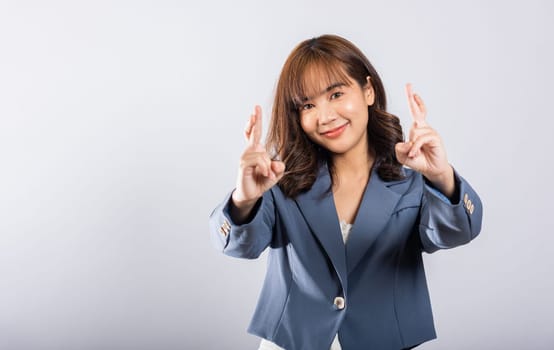 An Asian woman smiles confidently in a studio portrait, her fingers crossed as a sign of hope and good luck. Isolated on white, her expressive face conveys happiness and optimism.