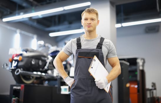 Portrait of qualified maintenance service worker in uniform, man with clipboard paper on motorbike background. Pit stop, garage, high quality help concept