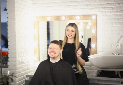 Portrait of pretty professional female master holding brush for modeling hairstyle. Happy male client in chair. Beauty studio, barber, barbershop concept