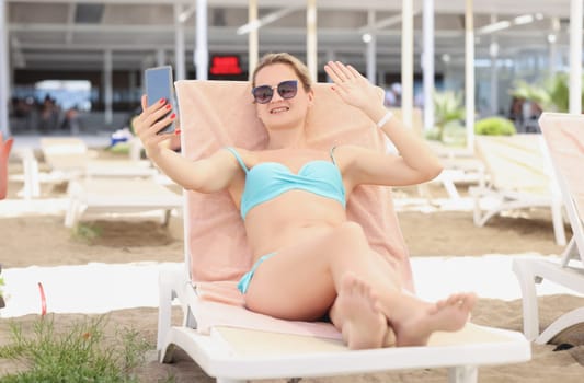 Portrait of young blonde woman lay on sunbed, wave hi to friend on video call. Pretty relaxed female in sunglasses. Summer, leisure, communication concept