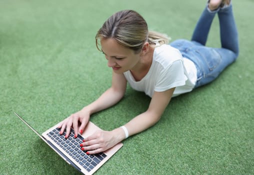Portrait of smiling blonde woman working on laptop lay on grass, remote job, female freelancer outdoors. Modern technology, communication, career concept