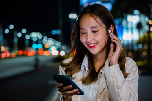 A portrait of a young woman talking on her smartphone with earphones, enjoying a conversation on a city street at night. Handheld device and wireless connection make it possible. Phone communication.