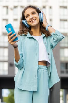 Happy relaxed overjoyed preteen child kid in wireless headphones choosing, listening favorite energetic disco rock n roll music dancing outdoors. Teenager girl walking in urban city street. Vertical