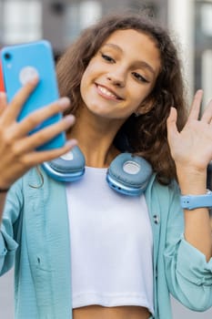 Pretty preteen child kid blogger taking selfie on smartphone, communicating video call online with subscribers recording stories for social media vlog. Teen girl walking in urban city street. Vertical