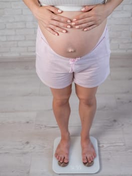 Top view of a pregnant woman in home clothes standing on an electronic scale and holding her hands on her tummy