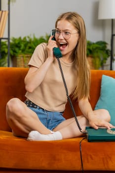 Young woman making wired telephone conversation with friends. Happy excited adult girl enjoying old-fashioned retro phone from 80s talking at home apartment living room sits on orange couch. Vertical