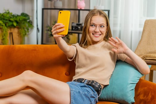 Young blonde woman blogger taking selfie on smartphone, communicating video call online with subscribers, recording stories for social media. Adult girl at home apartment living room sitting on sofa