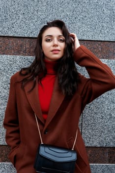 fashionable young woman in a brown coat and red sweater poses against the wall
