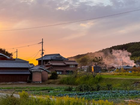 Smoke rises from fire by traditional houses in rural Japan with sunset glow in sky. High quality photo