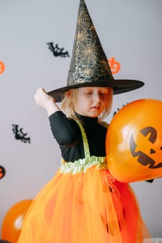 Children's Halloween - a girl in a witch hat and a carnival costume with airy orange and black balloons at home. Ready to celebrate Halloween.