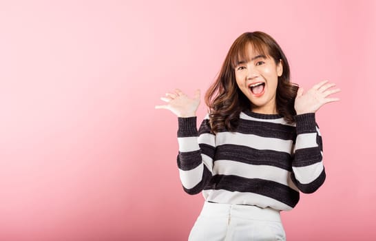 A smiling, happy woman raises her fists in celebration, gesturing success. Asian portrait of a beautiful young female expressing excitement and saying yes in a studio shot on a pink background.