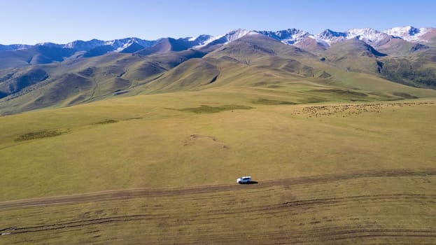 A white SUV is driving in the mountains through the fields. Aerial view from a drone of huge white clouds, snowy mountain peaks and green fields. Dirty off-road with puddles. Splashes from the wheels