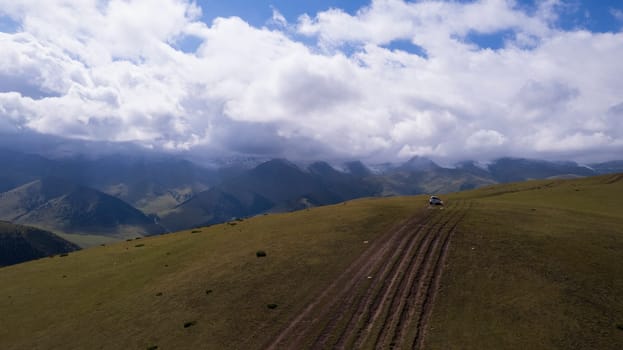 A white SUV is driving in the mountains through the fields. Aerial view from a drone of huge white clouds, snowy mountain peaks and green fields. Dirty off-road with puddles. Splashes from the wheels