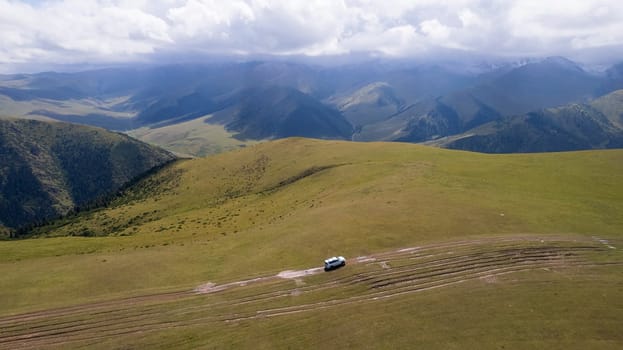 A white SUV is driving in the mountains through the fields. Aerial view from a drone of huge white clouds, snowy mountain peaks and green fields. Dirty off-road with puddles. Splashes from the wheels