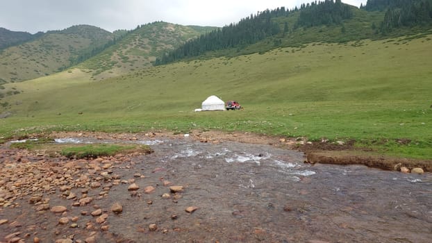 A white yurt among green fields and a river. Dark white clouds and fog lie on the green hills. A mountain river with stone banks. Horses graze in the distance. There is a red SUV next to the tent.