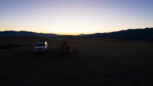 Sunrise among green fields and hills. Camping. There is a tent, there is smoke from the stove. There is a white SUV car nearby. The sun rays come out from behind the horizon. Nature of Kazakhstan
