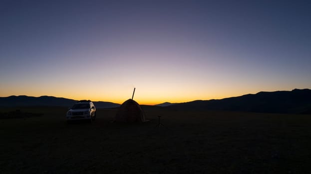 Sunrise among green fields and hills. Camping. There is a tent, there is smoke from the stove. There is a white SUV car nearby. The sun rays come out from behind the horizon. Nature of Kazakhstan