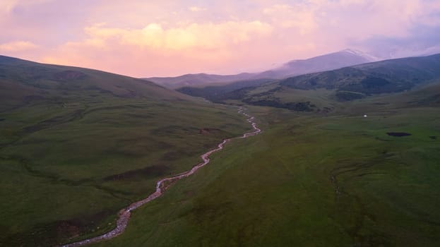 Pink-purple sunset in the mountains with green fields. There is a white yurt, a mountain river runs. Huge clouds. A herd of horses is walking. An SUV next to the mud and a tent. Tourists gather