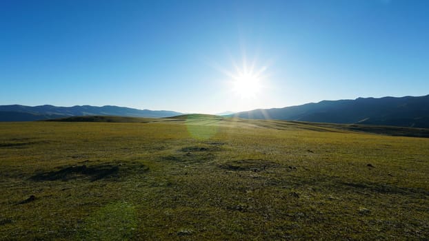 Sunrise with a view of green hills and snowy peaks. Spacious green fields and yellow grass in places. Blue clear sky. Shadows from mountains, gorges. Peace and relaxation. Outdoor recreation