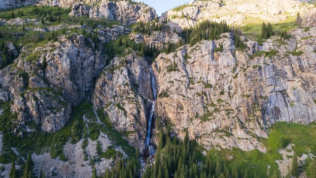 A high waterfall cascades in a mountain gorge. Coniferous trees, Christmas trees and grass grow. The river runs. Hiking trails. Top view from a drone. Sunset. Burkhan Bulak Waterfall, Kazakhstan