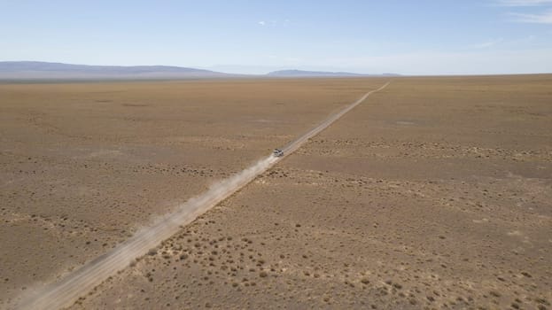 A white SUV is driving fast on a dusty road in the steppe. Clouds of dust fly from under the wheels and from the roof of the car. The jeep easily passes a dirt road. Blue sky and white clouds.