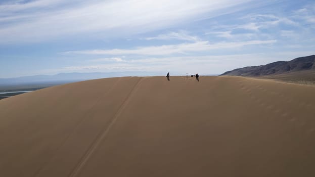 A large sand dune in the middle of the steppe. Drone view of a huge pile of sand. Tourists are walking, enjoying the view. A river runs in the distance and grass grows. The sky with white clouds