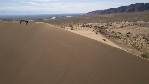 A large sand dune in the middle of the steppe. Drone view of a huge pile of sand. Tourists are walking, enjoying the view. A river runs in the distance and grass grows. The sky with white clouds