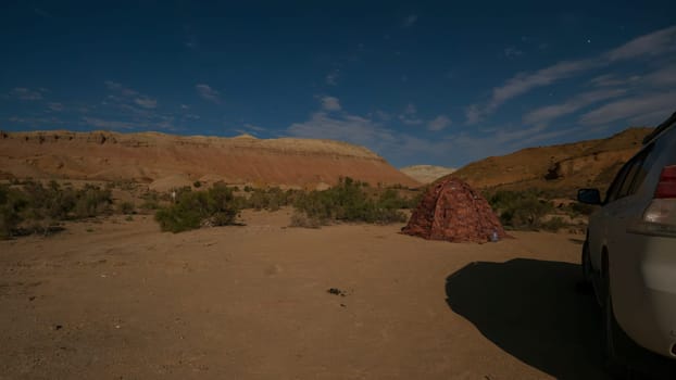 A bright starry night in the colored mountains. There is a tent and a white car. Stars and white clouds are visible. In the distance, colored limestone mountains. Green trees and bushes grow.