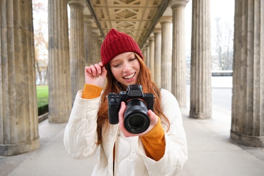 Young redhead female photographer, makes lifestyle shooting in city centre, takes photos and smiles, looks for perfect shot, makes picture.