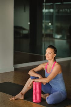 A slender girl is sitting on a yoga mat with a big massage roller in the fitness room.