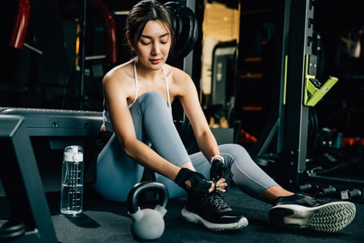 Woman tying her shoelace while sitting on the gym floor, holding a shaker bottle of water. The shot highlights her fitness and hydration routine.