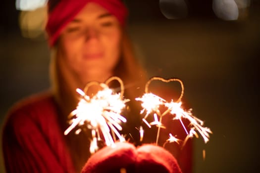 Woman holding sparkler night while celebrating Christmas outside. Dressed in a fur coat and a red headband. Blurred christmas decorations in the background. Selective focus.