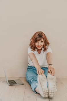 A brunette sits on the floor with a laptop on a beige wall background. She is wearing a white T-shirt, jeans and white sneakers