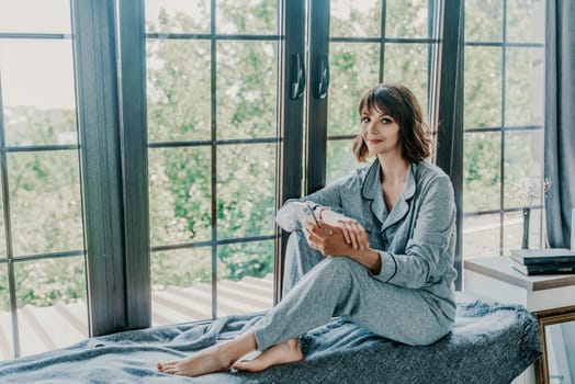 Woman window sill pajamas. Resting woman in pajamas. A calm girl with a phone sits on the windowsill at home. Side view.