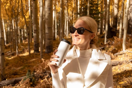 Beautiful Blonde Woman Drinks Hot Drink Coffee Or Tea From White Thermos Travel Mug In Autumn Park, Forest While Hiking. Relaxed Female In Her 30s Enjoys Camping, Breathing Fresh Air In Fall Season