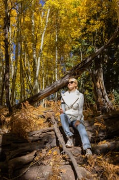 Pretty Woman Sits On Fallen Tree And Drinks Hot Beverage From Thermos Travel Mug In Forest, Nature, Wears Warm Clothes. Autumn Drink, Fall Leaves Season. Zero Waste. Vertical Plane. Copy Space.