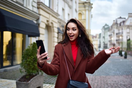 A young woman in a red coat walks down the street and experiences joyful emotions looking at the smartphone in her hand. Good News, Winning, Discounts