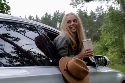 Blonde woman sticking head out of windshield car and drink coffee or tea from reusable mug. Young tourist explore local travel making candid real moments. Refuse reuse recycle zero waste concept. Responsible traveling reduce carbon footprint sustainable lifestyle