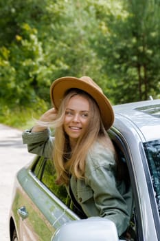 Smiling young woman in hat looking from car window. Local solo travel on weekends concept. Exited woman explore freedom outdoors in forest. Unity with nature lifestyle, rest recharge relaxation