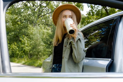 Blonde woman stoped on road next to car and drink coffee or tea from reusable mug. Refuse reuse recycle zero waste concept. Young tourist explore local travel making candid real moments. Responsible traveling reduce carbon footprint sustainable lifestyle