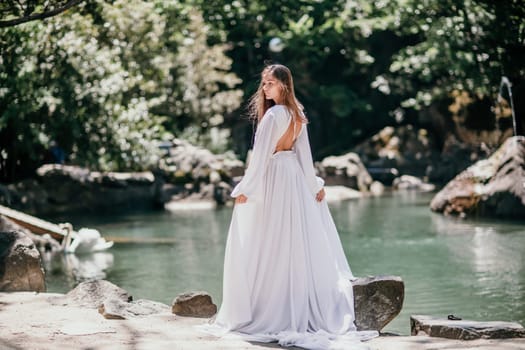 a beautiful woman in a long white dress looks into the distance at a beautiful lake with swans