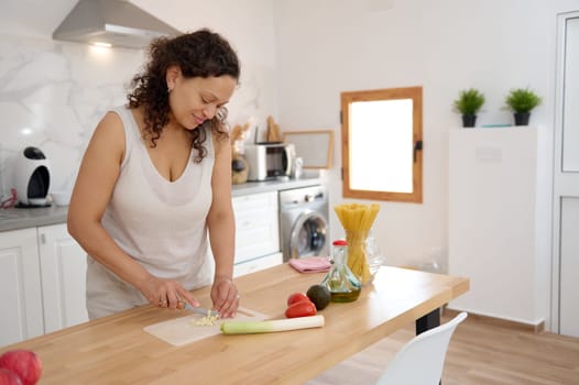 Young adult Latin American beautiful woman in casual clothes, cutting garlic, preparing a sauce for Italian pasta in the home kitchen. Healthy lifestyle, eating and dieting concept