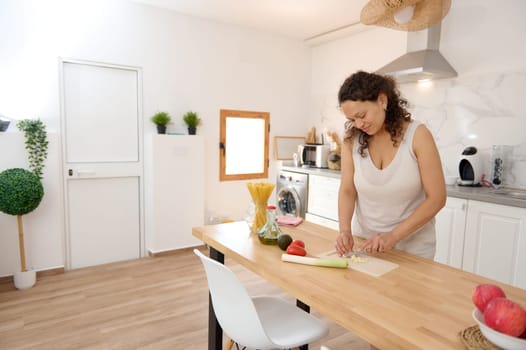 Beautiful young woman standing at modern kitchen, chopping vegetables, preparing fresh healthy salad for dinner or lunch. Young Latina woman cooking at home, making breakfast following healthy diet