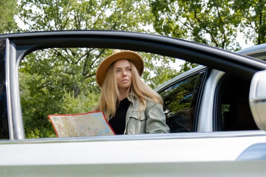 Smiling young woman in hat stoped on road to check the route on travel map. Local solo travel on weekends concept. Exited woman explore freedom outdoors in forest. Unity with nature lifestyle, rest recharge relaxation