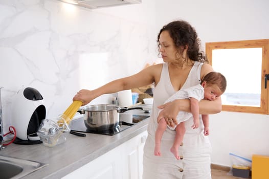 Happy Latin American woman, a loving mother, housewife holding her newborn baby, taking off spaghetti from glass container while cooking dinner in home kitchen. People. Family. Household and lifestyle